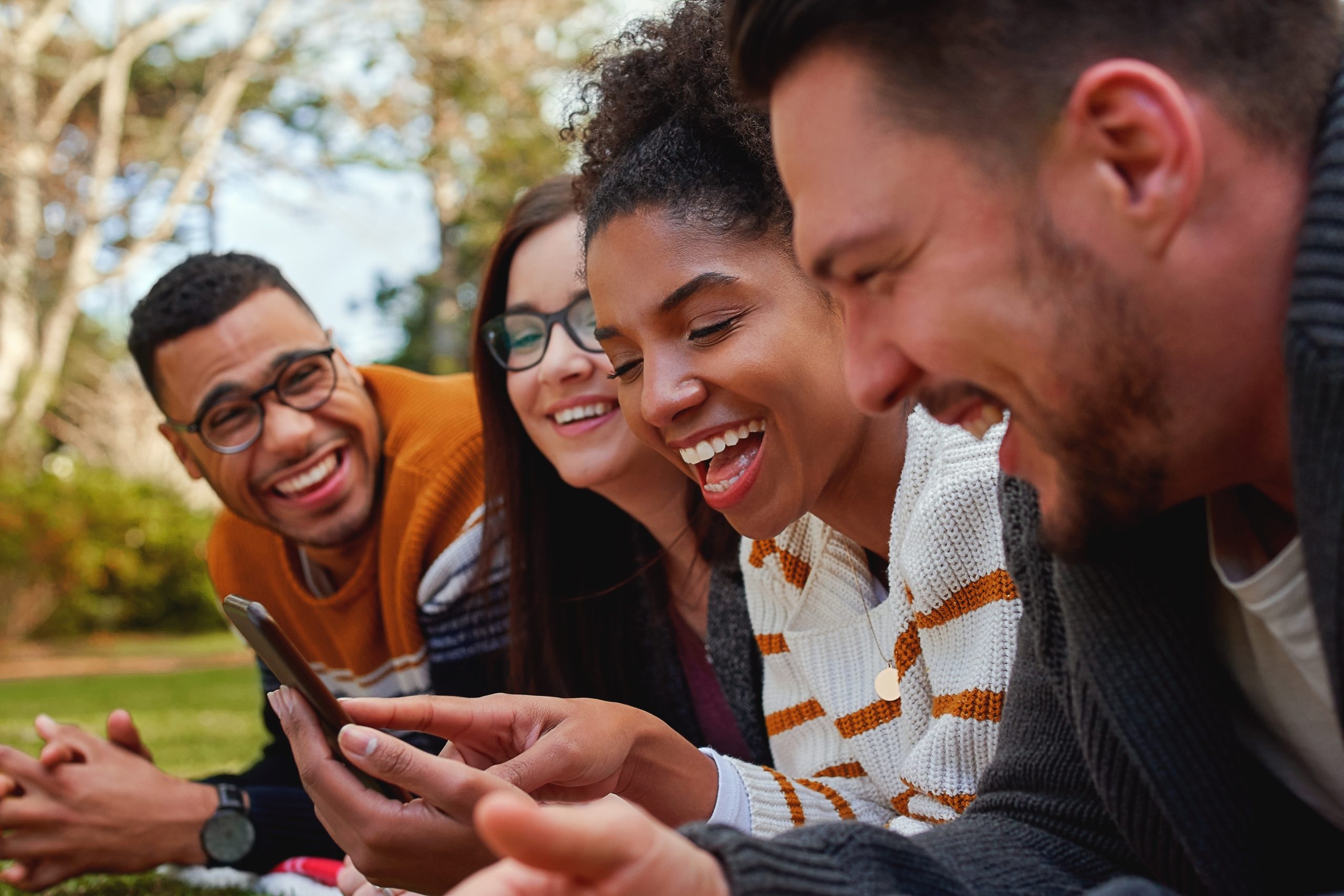 group of multi ethnic friends in college lying together in the park enjoying watching text or video on mobile phone - smiling group of students